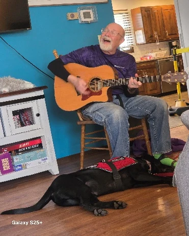 Hospice volunteer playing acoustic guitar and singing with his faithful therapy dog resting peacefully at his feet